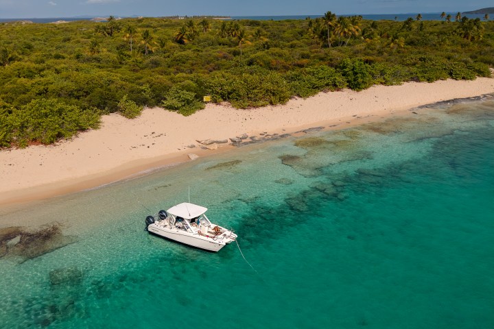 Boat anchored in clear turquoise water near sandy beach and lush green trees.