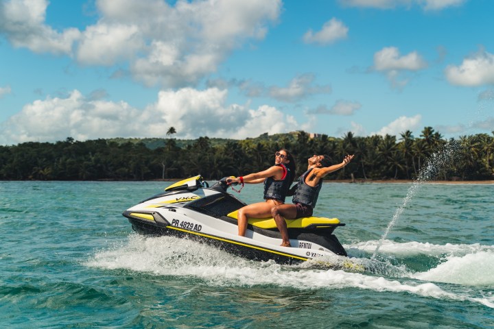 Two people on a jet ski in the ocean, green landscape in the background.
