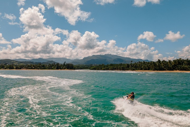 Person on a jet ski in turquoise water near a tropical shoreline with mountains and clouds.
