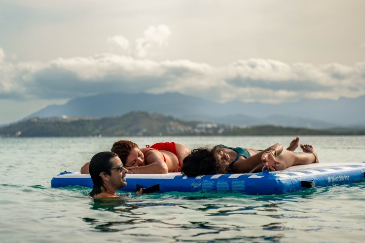 Four people relax on a floating mat in calm sea with mountains in the background.