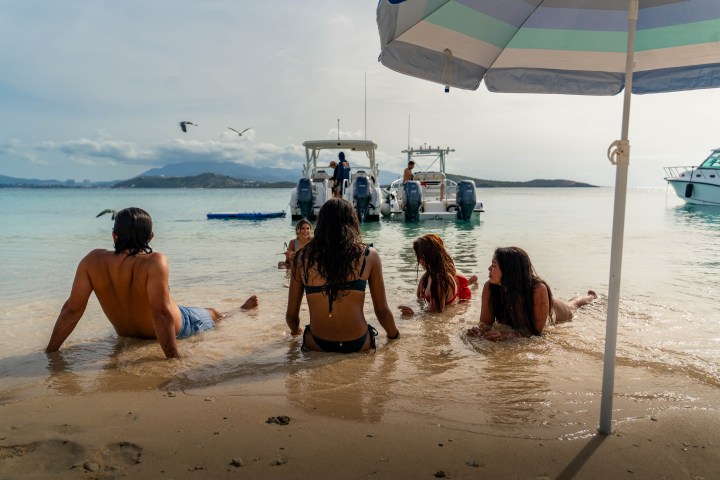 People relaxing on a sandy beach under an umbrella near boats.