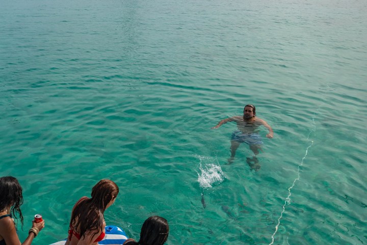 People on a boat watch a person swimming in clear blue water with a yacht and seagulls in the background.